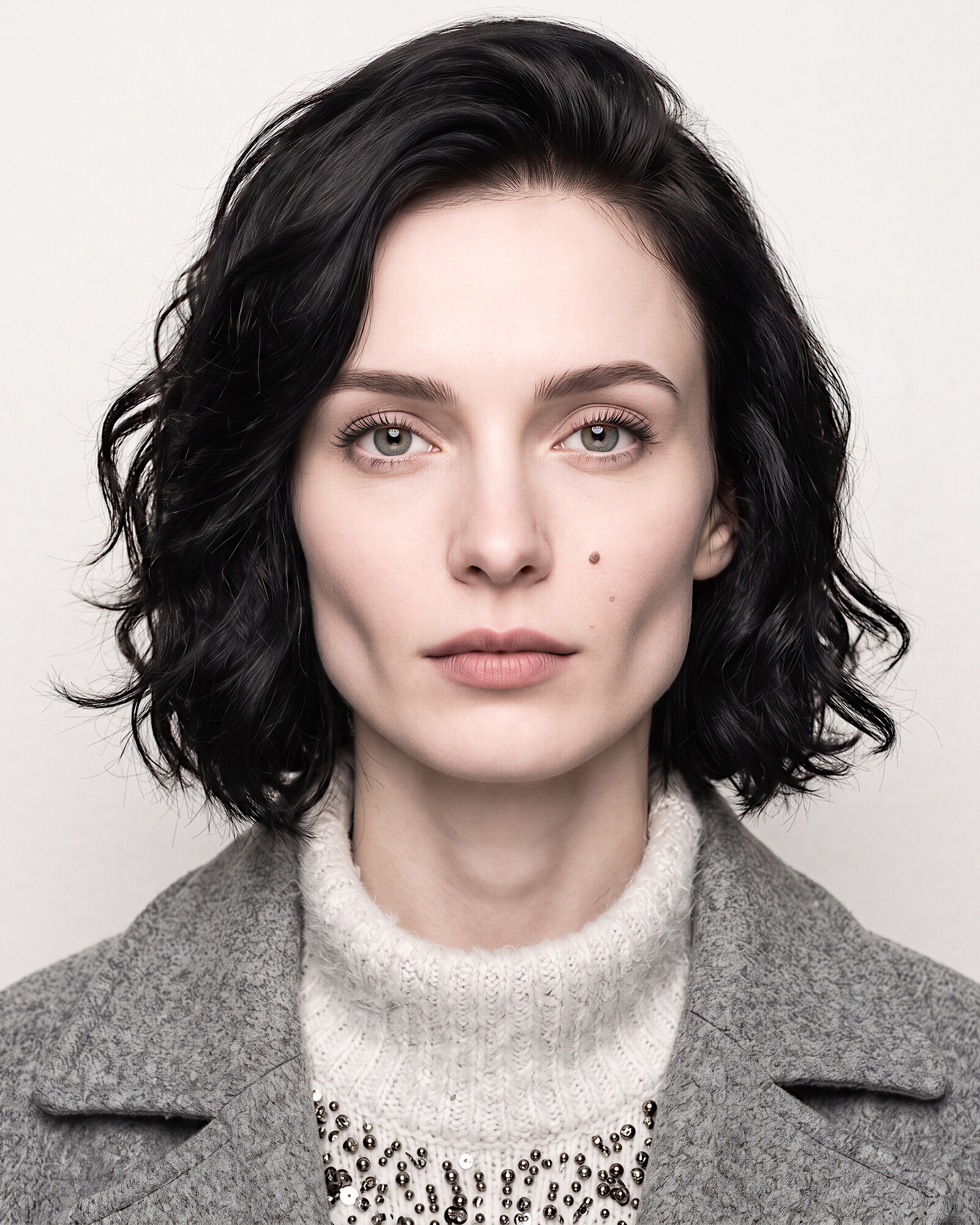A directed character — a black wavy bob, a grey wool coat over a white knit with beading at the neck